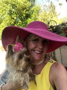 Smiling woman in yellow shirt and pink floppy hat holding dog. 